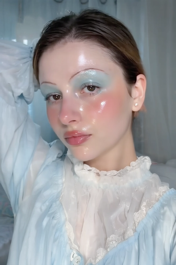 A close-up iPhone-style portrait of a young woman with a naturally soft hairstyle, her complexion glowing with dewy, glossy wet skin that reflects cool-toned lighting softly illuminating her face. Her eyeshadow is frosty pastel blue with pearlescent shimmer blending gently across her lids, complemented by heavy pink blush extended across her cheeks and beneath the eyes for a flushed babydoll effect. Her lips have a glossy, soft pink sheen that catches light in a tactile, natural way. Eyebrows are natural dark brown, brushed upward but unbleached, adding to the natural look. She wears a delicate babydoll-style blouse in pale icy blue with puffed sleeves and subtle lace trim around a high neckline, featuring sheer layers that softly diffuse the cool light. The image is framed tightly from just above her head to just below her collarbones, taken head-on with a slight casual tilt and relaxed expression, her gaze softly forward but not fully engaging the camera, adding to candid intimacy. The background is softly blurred in cool pastel tones with hints of indoor texture like a faint curtain or bedding. Lighting mixes soft daylight filtered through a window with subtle cool-toned phone flash, creating realistic shadows and gentle reflections on her wet skin and fabric textures, with detailed visibility of fine hairs, skin pores, gloss texture, and fabric weave, evoking a tangible, intimate spontaneity typical of authentic iPhone photography.