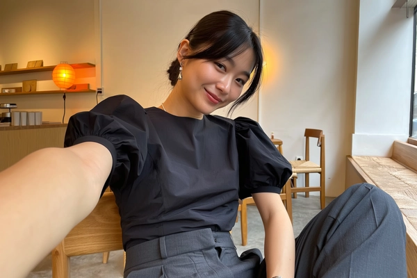 A spontaneously captured iPhone photo featuring a seated woman softly gazing into the camera inside a contemporary Japandi-inspired café. She wears a minimalist black puff-sleeve blouse neatly tucked into structured high-waisted trousers, embodying relaxed elegance with subtle texture contrast. Her understated silver jewelry adds a delicate touch of refinement. Soft natural daylight mixes with the warm ambient glow of an orange lamp in the background, illuminating the cafe's clean lines, warm wood furniture, and minimalist décor including simple shelving and muted textiles. The composition is casually framed with a gentle tilt, evoking warm, cinematic intimacy and authentic candidness. Visible details include realistic fabric grains, natural skin textures with subtle imperfections, loosely styled hair strands, and softly diffused shadows enhancing the genuine iPhone snapshot feel.