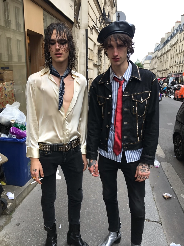 Two ultra-slim young men stand side by side on a Parisian sidewalk, framed in a spontaneous mid-shot with a casual, slightly off-center crop that catches them in a natural, unposed moment. One wears a silky cream shirt unbuttoned halfway down, revealing a slender chest and a loosely knotted skinny scarf, paired with a studded leather belt clinging to black skinny jeans. Classic black oxford shoes peek beneath worn hems. His long, shaggy hair looks damp and unkempt, obscuring tired eyes rimmed with smudged black eyeliner. A cigarette is loosely held in his right hand, fingers relaxed. The second wears a black denim jacket heavy with gold studs, layered over a faded striped shirt and a loosely tied red skinny tie. Metallic silver ankle boots catch uneven daylight. His black leather beret sits slightly askew atop messy hair with visible roots. Both have a skin texture that reveals subtle pores, faint redness, and authentic imperfections. Tattoos pepper their hands, lending raw edge. Behind them, a gritty Paris street scene unfolds—Haussmann buildings with chipped stone facades and overflowing trash bins. The daylight is muted and diffuse, casting soft, natural shadows accented by the slight desaturation typical of an unfiltered iPhone snap. The image radiates a cool, exhausted, post-party energy, a snapshot of stylish chaos and indie sleaze glam caught just after a frenetic fashion event, intimate and immediate.