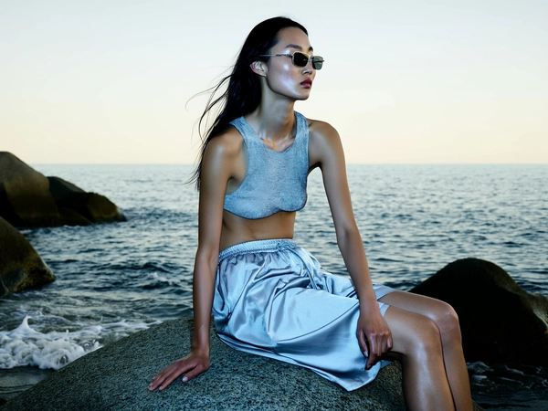 A digital flash softly illuminates an East Asian woman seated on a rugged rock, her cropped sleeveless top revealing subtle skin textures, the oversized pants pooling like satin folds around her legs. She grips her hair back, a quiet tension in her pose that balances stillness and the restless sea breeze. Her angular sunglasses catch the flash’s glow, faintly pulsing with cool luminescence against calm ocean waters that retreat into a slightly overexposed horizon.

The setting is a minimalist natural stage: the matte grey stone she rests upon contrasts with the creamy softness of the sea and sky, a restrained palette enhanced by the cool digital flash and gentle ambient light. The fabric weave of her top is palpable under close scrutiny, each thread casting micro-shadows that deepen the garment’s tactility. Fine skin pores along her jawline and collarbone catch shards of light, highlighting a fresh, natural complexion.

Framed from a mid-height vantage point with a 85mm lens, the composition isolates the model against the vastness of the sea while preserving intimate detail. A slight telephoto compression flattens the distance, while a narrow depth of field renders the rock’s texture crisply, letting the seascape dissolve into luminous softness behind her. The image feels quiet yet charged, poised between stillness and a halting gust of wind.

The visual tone echoes subtle digital clarity combined with an ultra-fine grain reminiscent of translucent film stocks, lending an almost painterly coolness without sacrificing the hyper-real texture fidelity of flesh and fabric—high-fashion editorial, hyper-real texture fidelity