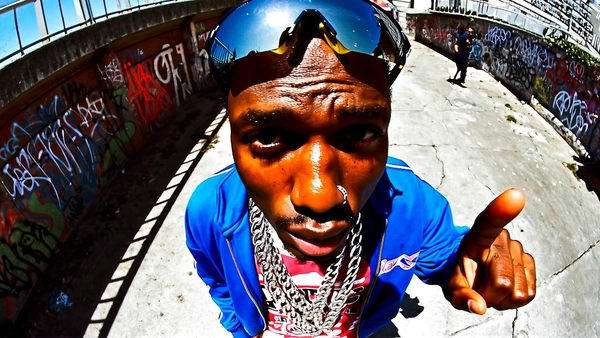 A wide shot of a Black man with warm brown skin and coarse natural skin texture visible across his forehead, nose, and cheeks, captured in a middle close-up from a top-down wide-angle perspective. He wears a bright cobalt track jacket zipped halfway over a bold graphic tee, heavy silver Cuban link chains draped prominently around his neck and large reflective sunglasses pushed up on his forehead. His face is close to the lens and slightly off-center, the fisheye distortion enlarging his nose, lips, and extended hand pointing assertively toward the camera.

He stands against an urban backdrop of cracked concrete with graffiti-tagged walls curving inward through the lens's warp. Overhead sunlight casts sharp shadows beneath his chin and accentuates the metallic shine on his chains. The image contains a subtle grain overlay enhancing skin texture sharpness, fabric creasing, and environmental surface detail. early-2000s Y2K snapshot