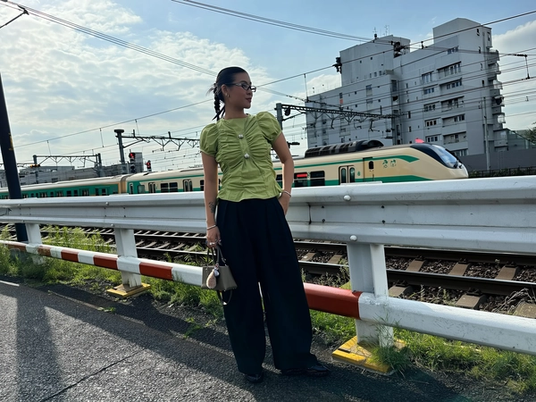 A young woman stands confident beside a closed suburban Tokyo railway crossing, illuminated by soft natural light spilling gently through scattered clouds. Her light olive-green ruched cap-sleeve blouse tucks effortlessly into wide black balloon trousers, while chunky silver rings and sleek glasses add cool edge. A tiny plush charm dangles from her mini handbag, its subtle leather grain catching faint whispers of midday glow. Slicked-back hair and faint arm tattoos peek in rhythmic contrast to the weathered railings and wild grass edging the uneven sidewalk beneath her feet. Behind, a cream and blue commuter train glides through overhead wires and faded apartment balconies, traffic lights blinking softly into a distant, subdued urban hum. The frame tilts slightly from a casual hip-height grab, edges cropping into textured asphalt and blurred wire silhouettes, evoking an off-guard moment held in tranquil pause—Tokyo street snap, shot on iPhone