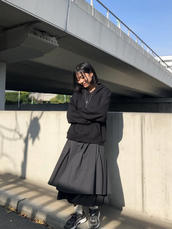 A spontaneously candid iPhone photo showing a young woman standing beside a minimalist concrete overpass under soft natural daylight. She wears a black oversized hoodie layered effortlessly over an architectural full skirt made of matte cotton fabric, its folded layers softly textured and flowing naturally. On her feet, she sports heavy, laced sneakers that add practical, grounded contrast to her elegant yet relaxed outfit. Above her, the overpass casts geometric shadow patterns onto the rough concrete wall, creating a visually compelling interplay of light and shadow. The scene is framed with a slight casual tilt, emphasizing the genuine spontaneous snapshot aesthetic, capturing realistic skin textures, natural hair strands loosely framing her face, and authentic environmental textures of concrete and fabric. The composition highlights Japandi-inspired minimalism with functional urban surroundings and thoughtful layering of minimalist garments.