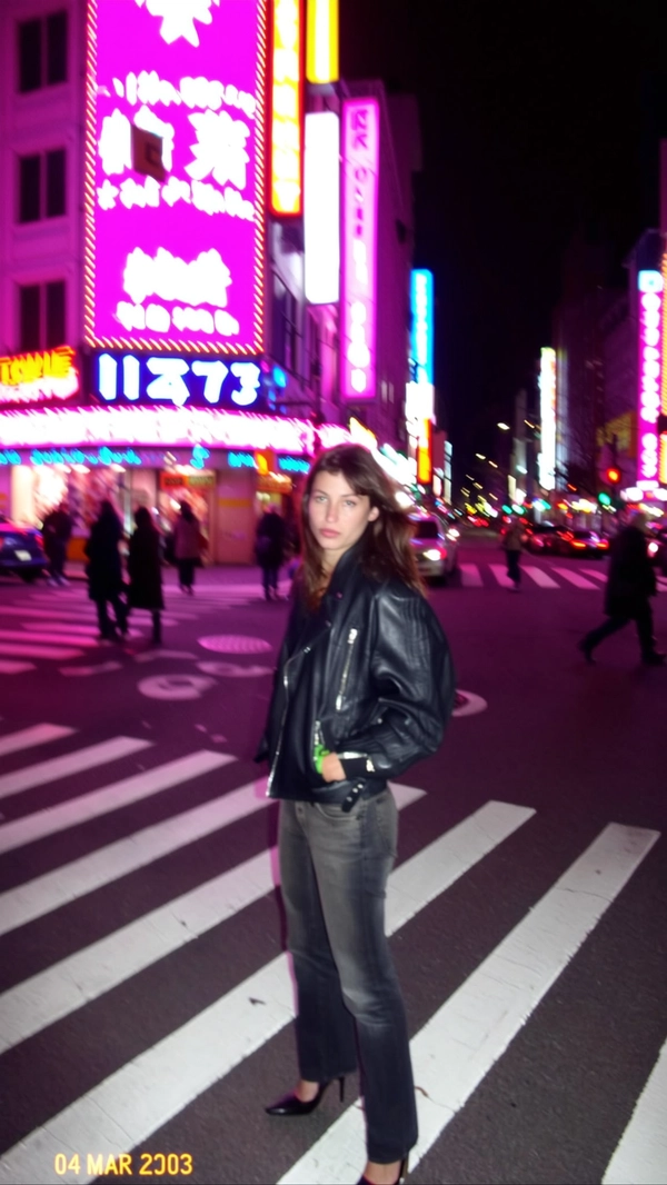 A sudden pop from the built-in flash cuts through the dim city street as a woman in a leather jacket and heels stands stiff at the pedestrian crossing, neon shop signs swirling behind her in a blur of pastel pink and blue glows. The CCD sensor struggles in the low light, speckling the shadows with grain and pixel fringing while the soft focus edges smear the glow into a hazy haze. The camera's shaky Dutch tilt adds to the restless tension in the frame, a jagged timestamp reading “04 MAR 2003  23:18” stamped in the corner next to a flickering battery icon. She blinks against the flash; the low-res capture feels like a stolen fragment from a late-night wander. The photo quality suggests a slightly grainy or low-resolution digital look, capturing a candid moment.—early-2000s digicam snapshot, grainy low-res quality