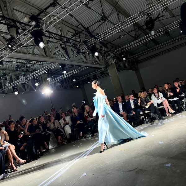 A striking model in a sharply sculpted icy blue dress commands the runway, the architectural folds of the fabric catching faint light and casting ethereal shadows over the raw concrete floor. She strides confidently past a front row packed with fashion editors and influencers, some mid-whisper, others lifting phones to capture the look. The venue’s industrial bones reveal exposed steel beams crisscrossing overhead, hung with cluster arrays of tungsten and cool daylight-LEDs that scatter glints over the dress’s subtle shimmer. Motion blurs trail from the dress’s angular protrusions and the crisp edges of patent leather heels, while taped sight-lines and occasional scuff marks punctuate the gritty walkway. The iPhone’s sensor breathes with slight digital noise in the shadow zones and blooms softly around intense spotlights, lending authentic handheld texture to the shot. Framed from an aisle angle just behind the seated row, the image tilts gently, capturing arrested movement and audience energies in a slice of live show magic—contemporary runway snapshot, shot on iPhone