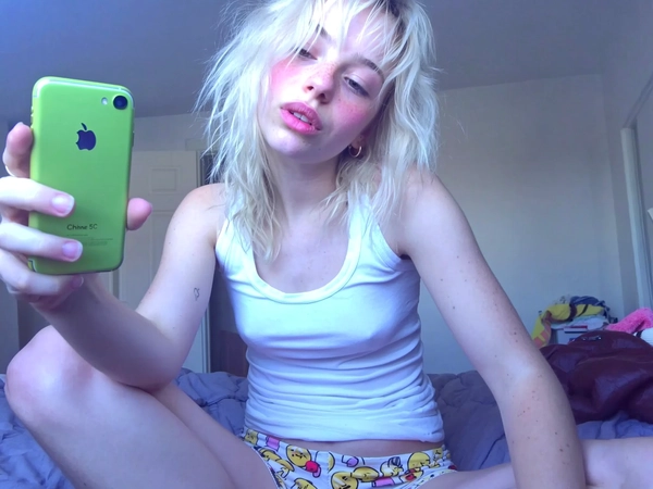 A candid indoor snapshot of a young woman with white-blonde, loosely tousled hair sitting on a bed wearing a simple white tank top and pastel emoji-printed shorts, holding up a green iPhone 5C. Her relaxed, slightly blank expression is visible with subtle, natural makeup including faint pink blush tones, while her skin shows slight overexposure consistent with natural ambient light filtering into a cozy bedroom. The image carries a distinct mid-2010s lo-fi aesthetic with intentionally muted color saturation, softened contrast, and cool-toned bluish-violet shadows contributing to a gently fading, analog-like digital softness. Visible textures include delicate fabric folds in cotton shorts, a slight sheen on the tank top, natural skin imperfections, and clearly defined, loose hair strands. The framing is casually off-center with a slight tilt, capturing an intimate, informal moment evocative of authentic mid-2010s candid smartphone photography.