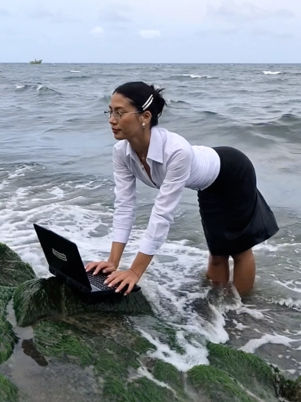 With waves lapping rhythmically against the shore, a businesswoman stands ankle-deep in seawater, her crisp white blouse and fitted black skirt sharply contrasting with the slick, moss-covered rocks beneath her. She leans confidently over the wet stones, fingers resting on an awkwardly propped laptop whose screen reflects the cloudy sky above. The photo has a grainy quality, suggesting it may be a still from a video or taken with an older digital camera. Her hair is neatly clipped back with silver barrettes, glasses perched on her nose, her expression one of composed determination as gentle waves wash around her calves. Quick snapshots create a soft-focus effect at the edges, and muted colors swirl into a nostalgic haze, capturing an absurd twist on remote work culture. The ocean’s unpredictable rhythm seems to pulse beneath the steady click of keys, while a faint breeze tousles the hem of her skirt—early-2000s digicam snapshot, grainy quality.