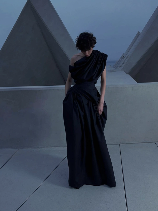 A softly dim-lit dusk-inspired iPhone snapshot featuring a model in an avant-garde floor-length black gown by Rick Owens, showcasing structured sculptural draping and an asymmetrical neckline with gathered fabric layered over one shoulder. The model stands slightly off-center against angular Brutalist concrete architecture under muted cool dusk hues of greys and shadowy blacks. Soft, diffused ambient twilight lighting accentuates the matte textures of the gown and subtle imperfections of the concrete surfaces. The composition employs a gently elevated viewpoint tilted downward, casually framing the model within expansive negative space, evoking relaxed asymmetry, quiet introspection, and intimate moody minimalism in a muted twilight palette.