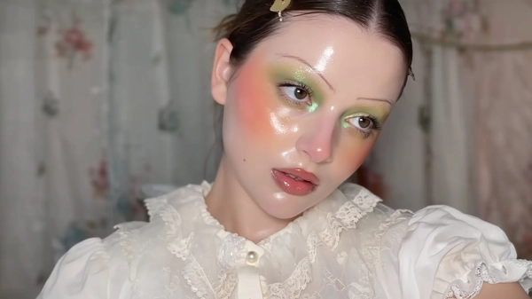 A close-up iPhone-style portrait of a glamorous young woman with glassy, dewy skin featuring visible pores and subtle natural texture. Her makeup showcases an iridescent blend of shimmering green and warm gold eyeshadows softly diffused across her eyelids, extending gently beneath her eyes in a babydoll style with a delicate glow. Her cheeks are flushed with a heavy, soft pink blush that spreads below her eye area, harmonizing with her glossy lips tinted in a soft peachy-pink hue. Her natural eyebrows are light brown, brushed upward with fine individual hairs visible, unbleached. She wears a vintage-inspired babydoll blouse in a pale cream tone with puffed sleeves and lace collar details, featuring tiny pearl buttons and a sheer, translucent overlay that softly captures the light. The photo is tightly framed from just above her head down to her collarbones, taken at a slight casual tilt to mimic a handheld iPhone shot. Her expression is relaxed and slightly dreamy, her gaze off-camera with parted lips suggesting spontaneity. The softly blurred background carries muted pastel tones with hints of indoor textures, creating a gentle, intimate atmosphere. Lighting is a natural mix of soft daylight and subtle iPhone flash, casting believable shadows and realistic highlights on her face and fabric textures. Fine details such as stray hairs, shimmer particles on the eyeshadow, fabric weave, and skin texture are crisply visible, producing a tactile, authentic image reminiscent of a candid iPhone portrait.