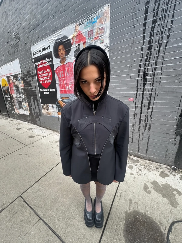A sharply stylized wide-angle iPhone photograph captures a woman standing confidently on a subtly wet urban sidewalk against a textured gray brick wall marked by streaks of rain and layered torn posters. The elevated viewpoint dramatically exaggerates her upper body proportions and visually softens her lower half. She looks directly up into the camera with a genuinely neutral, slightly intense gaze and relaxed posture. Her outfit is an original reinterpretation inspired by sculptural outerwear featuring asymmetrical paneling and matte industrial-look fastenings, paired with semi-opaque tights subtly patterned with delicate woven textures, and chunky platform shoes crafted from contrasting mixed materials. Her hair is styled in a sleek, contemporary cut framed softly to balance the architectural feel of her coat. The natural lighting is a softly diffused overcast daylight filtering urban skies, casting nuanced shadows that subtly reveal fabric textures and urban wall imperfections. The composition embraces authentic wide-angle distortion and casual iPhone framing, highlighting the subject's poised presence and awarding the scene a dynamic yet effortlessly stylized street fashion portrait vibe.