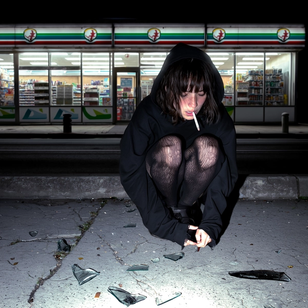 A young woman perches on chipped concrete, her loose hoodie draping in shadowed folds, torn tights revealing fleeting glimpses of skin textured with urban grit. Her messy bangs slip over eyes caught mid-blink, a cigarette smoldering between slender fingers, embers flickering against pale skin pores and faint freckles. The convenience store behind blurs into a stark, washed-out tableau, flattened by the harsh, direct flash that splinters from the cracked sidewalk beneath.

The flash carves the scene into layers of harsh white light and deep black shadows, the cigarette’s smoke curling in crystalline detail against the night. Subtle hints of residual neon flicker and refract softly on the cracked pavement, its granular surface and scattered glass shards absorbing and refracting splinters of cold light. Her hoodie’s worn cotton weave and the tights’ frayed mesh pull focal attention to tactile imperfection.

Captured with a 50mm standard lens at eye level, the composition centers her silhouette, cropped tightly to emphasize urban solitude and vulnerability. The flattened backdrop contrasts with the textured foreground, evoking a raw intimacy within a gritty nocturnal world.

Digitally rendered with an analog-inspired high-contrast profile that blends stark flash realism and muted color saturation—high-fashion editorial, hyper-real texture fidelity