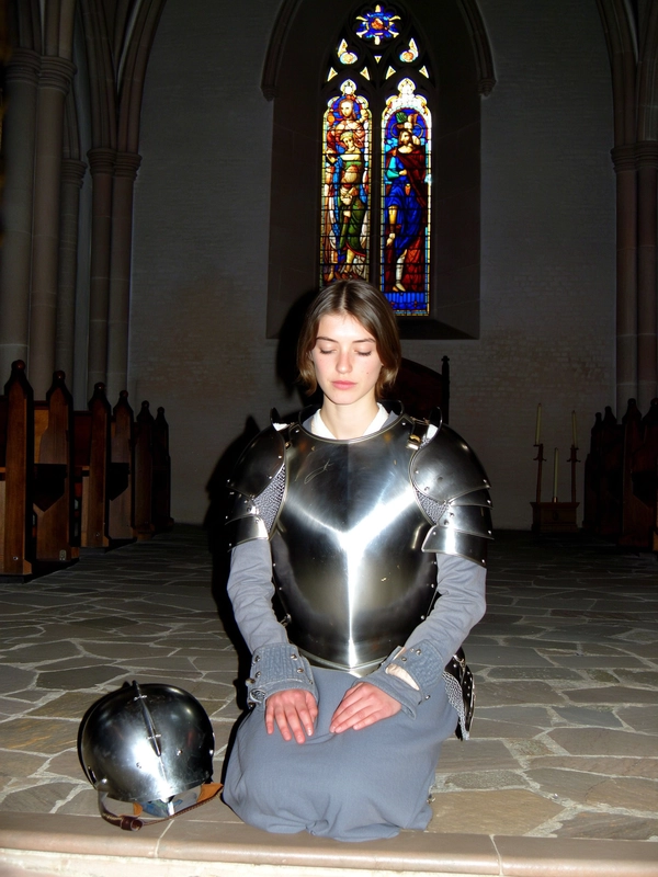 A wide, full-body, eye-level shot from an early 2000s-style digital camera explicitly capturing a young woman kneeling gracefully on one knee inside a dimly lit medieval church before a stained glass window. She wears battle-worn, partial medieval armor with visible subtle scratches and patina, her helmet placed neatly beside her on the stone floor. Her posture is flawless and solemn, hands resting gently on her thighs, eyes closed in calm reflection. The dim ambient light combined with harsh direct flash creates subtle, colorful reflections from the stained glass window dancing softly on her metal armor, emphasizing authentic worn textures and fabric details. The composition is thoughtfully balanced and elegant, highlighting her introspective knightly refinement within the high-contrast, gritty early 2000s digital photographic aesthetic blending medieval-core fashion and detailed realism.