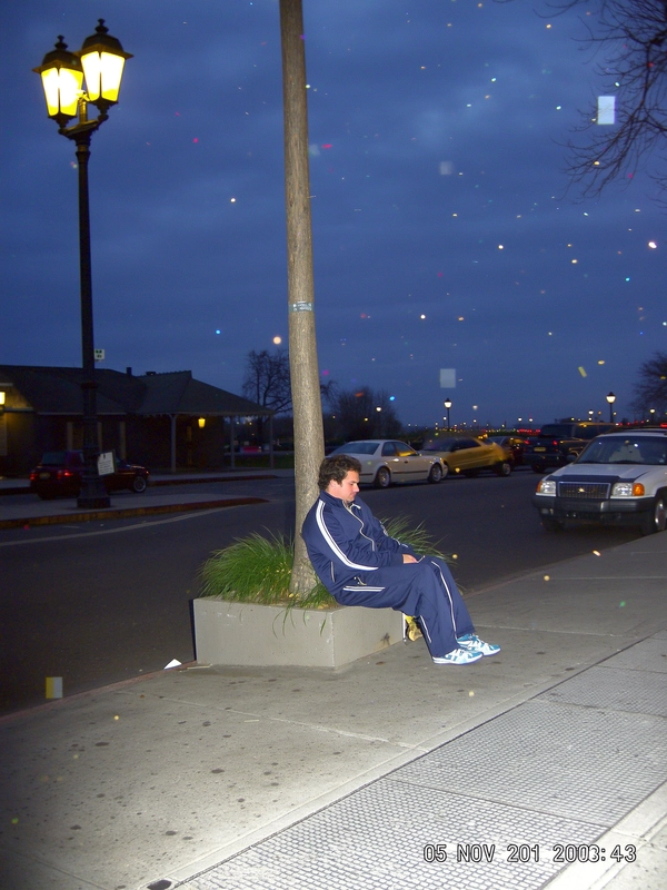 The soft glow of a late evening streetlamp washes over a quiet sidewalk where a man lounges on the edge of a concrete planter, his crisp tracksuit slightly wrinkled at the elbows as he slouches with easy intent. The on-camera flash pops suddenly, casting harsh shadows behind him and scattering pixel fringing across the dim pavement. Around the edges, CCD noise speckles dance like dust in a lazy breeze, while faint JPEG blocks blur the outlines of distant cars parked mid-frame. The composition is cocked at a Dutch tilt, making the lantern light skew the colours into muted blues and mustard yellows. Stamped faintly at the bottom-right, "05 NOV 2001  20:43" marks the moment, with a blinking battery icon striving for life at top corner. The photo quality suggests a slightly grainy or low-resolution digital look, capturing a candid moment.—early-2000s digicam snapshot, grainy low-res quality