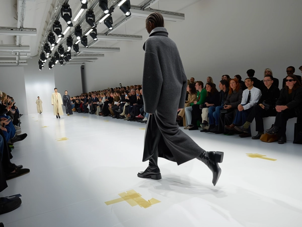 A poised model cloaked in a sculpted head-to-toe ensemble of tactile wool layers and sleek black leather boots strides onto a pristine, glossy white runway, the fabric’s matte depth contrasting with the subtle sheen of leather panels. Her sharp heels click deliberately, sending a focused rhythm into the hushed room as dozens of eyes fixate from benches running parallel, some guests leaning forward mid-whisper. The minimalist venue frames her with stark white walls and a harsh linear glow from overhead fluorescent tubes mingled with angular spotlights, casting soft shadows that ripple along her silhouette. The iPhone’s sensor catches the soft wool weave’s fine hairs softly illuminated, slight motion blur clutching the second heel in mid-air while the leather catches glints of ambient lighting. Gritty taped guidelines and light scuffs mar the glossy runway surface, anchoring the spontaneity. The viewpoint angles slightly from aisle-level, the slight hand shake lending intimacy to this poised entry—contemporary runway snapshot, shot on iPhone