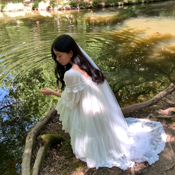 A quietly poetic moment gently captured on an iPhone, presenting a black-haired woman kneeling beside a serene forest clearing pond, adorned in a flowing white Jenny Fax dress crafted from sheer fabric with intricate lace trims and delicate layered ruffles. A long, softly trailing veil embroidered with subtle floral motifs cascades gracefully behind her. Her hair is softly styled with natural waves framing a quietly introspective, fresh-faced expression with minimal makeup enhancing her gentle femininity. Soft dappled sunlight filters through overhead leaves, casting warm, gentle patterns across the lace detailing and the shimmering water surface. The natural setting features subtly textured moss and weathered tree bark, complementing the tender vintage atmosphere. Framed with a slight tilt and elevated viewpoint, the composition balances spontaneous intimacy with whimsical delicacy, highlighting the iridescent fabric layers, the delicate embroidery, and peaceful reflective water, all suffused with a soft pastel mood emblematic of Jenny Fax's ethereal style.