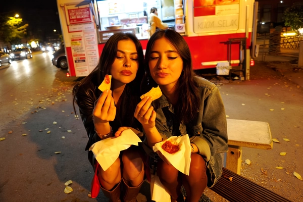 Beneath the dim amber haze of a lingering streetlight, two best friends lean close, illuminated by the neon pulse of a lively food truck. Their sleek hair, slightly tousled from the night, frames faces marked by the soft smudge of midnight eyeliner, lips bare but for a trace of natural gloss. Both clad in effortlessly cool off-duty chic—casual jackets draped over bare ankles, heels discarded in the gravel beside them—embody an unpolished allure.

One friend lifts a crisp tortilla chip, laden with melting cheese and a dot of vibrant salsa, poised with precise calm before the first bite. Their expressions hold steady, eyes half-lidded yet serene, lips parting delicately around the savory morsel. The crunch seems almost audible in the stillness, contrasting with the sleepiness in their shared gaze.

Around them, the urban backdrop softens into blurred streaks of color and shapes: a worn bench, scattered leaves, the flicker of passing car headlights. The textures are tangible—gritty asphalt underfoot, glossy napkins catching stray drips of salsa, the rough weave of a denim jacket. This quiet pause breathes authenticity and ease.

Captured from a low, arm’s-length angle that tilts slightly off-center, the composition feels intimate and spontaneous. Warm streetlight casts soft shadows, bathing the scene in mellow gold hues reminiscent of early dawn solitude. The moment flushes with subtle energy—laid-back glamour entwined with the familiarity of long friendship, perfectly resembling a candid iPhone photo.