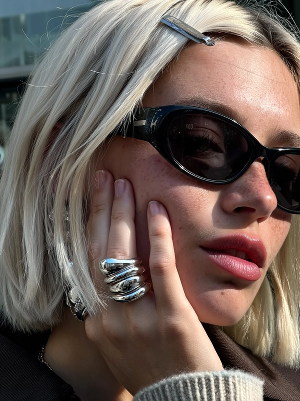 A platinum bob slips loose from a claw-clip, framing a woman’s face with frosted lips and a smudge of sunlit freckles. Her hand presses lightly to her cheek, stacked with chunky silver rings that glint like molten metal, bending in fluid, bold shapes. Black wrap-around sunglasses catch the faint reflection of city glass and the phone’s screen, while cool overcast light throws soft shadows on skin with pores and the fuzz of a wool rib cuff nearby. The shot tilts just off-center, fall-off blur wrapping around her cheek and shading, capturing the casual disarray of an artful moment cropped tight—close-up captured on Iphone, hand-face jewelry focus