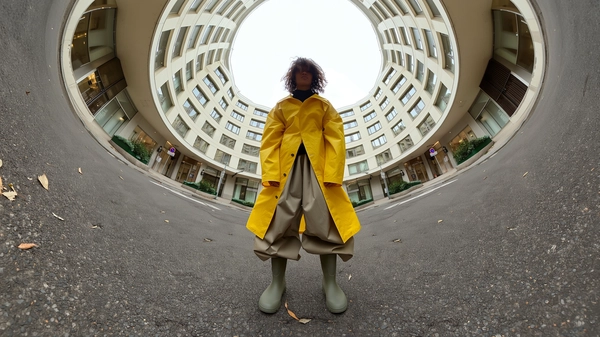 A spontaneous stylish 360-degree candid image captured from ground level in the middle of a quiet urban roundabout under a softly curved dome of surrounding buildings. A solitary figure stands naturally center frame, wearing an effortlessly chic sculptural yellow raincoat crafted from glossy, textured waterproof fabric, paired with massive, wide-flared pants in a complementary neutral tone with subtle drapery and soft folds. The figure's posture is relaxed yet confident, arms hanging loosely, head slightly tilted, hair tousled naturally with nuanced texture and light imperfection. The footwear consists of matte-finish rubber boots with gentle scuff marks typical of everyday wear. The ambient lighting is a diffused overcast daylight casting soft, even illumination, gently accentuating the smooth architectural surfaces of the curving buildings forming a seamless circular dome overhead. The ground is a finely textured asphalt surface with subtle cracks and scattered leaves, adding to authentic urban tactile realism. The spherical, circular upward-facing composition from the central floor-level viewpoint creates an immersive tunnel-like effect, enveloping the viewer within the dome and distorting perspectives with elegant visual curvature characteristic of spontaneous 360-degree photography captured by a portable camera connected to an iPhone, emphasizing the figure's stylish presence with natural candid spontaneity and detailed fabric and environmental textures.