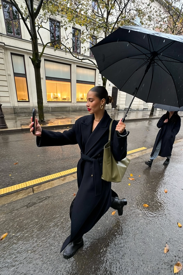 She strides beneath an overcast sky on slick pavement, long black coat buttoned sleekly over an olive shoulder bag, her low bun neat with glistening wet strands, hoop earrings catching muted light and lips bright with red that pops gently against the subdued palette. Her right arm extends forward holding a large black umbrella and the iPhone for a candid selfie, the lens angled wide capturing wet stone façades, sparse-leafed trees framing the street, and another woman a step behind also cloaked in dark layers and an umbrella canopy. The slick sidewalk reflects a whisper of golden sheen from the damp pavement, umbrella ribs texture visible against dim daylight, and the neutral city block's large windows glow faintly. Slight tilt invites imbalance and immediacy, while fine raindrops bead on umbrella fabric and lens edges, motion blur hinting at her step—rainy city snap, shot on iPhone