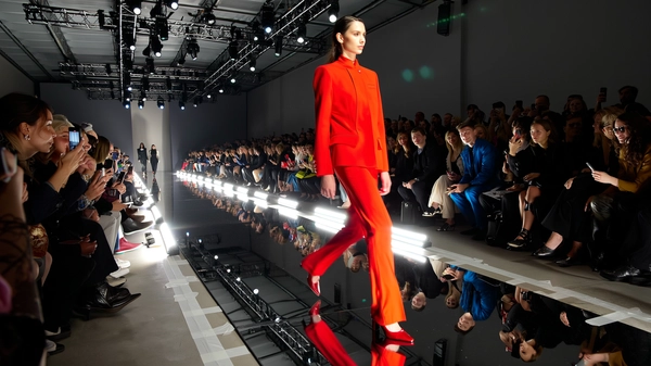 A statuesque model in a sharply tailored, vivid red suit commands the runway, the long-legged silhouette striking against a high-gloss black runway that mirrors her every confident step. The jacket’s crisp lines and slight sheen bounce catching flickers from overhead tungsten and LED spotlights, while sleek leather pointed heels blur with subtle motion as she strides solo. Reflected light stretches beneath her feet, doubling the intensity of the saturated scarlet tone. The venue’s minimalist industrial architecture frames the scene, with rigging bars and glowing gelled PAR cans visible overhead. Around the runway, the front-row crowd leans forward, phones raised, some whispering, others with captivated faces caught in warm bounce light. The iPhone’s sensor softens the contrast slightly, rendering gentle halos around bright reflections and bringing a just-noticeable digital grain to dimmer areas. The concrete floor nearby shows taped runway markers scuffed from earlier rounds. Shot from an off-centre aisle angle with a subtle handheld tilt, this snap vibrates with immediacy and editorial confidence—contemporary runway snapshot, shot on iPhone