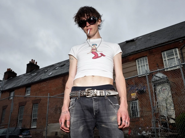 A skinny young man stands in front of a weathered brick building and a graffiti-tagged chain-link fence, framed from a sharp low angle that slices the frame unevenly, evoking the grainy immediacy of an old iPhone 3G snapshot. Overcast gray skies cast a flat, muted light, washing out colors except for his pale, unfiltered skin and the tight, cropped white tee that clings to his torso, riding up to reveal hip bones and a faintly mottled texture beneath. His faded shirt sports a distressed red-white-blue mermaid graphic, the cotton threadbare and stretched unevenly around the neckline. A cracked, double-pronged studded belt holds his low-rise, slightly slouched faded black jeans, frayed at the pockets, with subtle dirt stains near the cuffs.

Perched crookedly on his face are large, chunky black sunglasses that obscure his tired, unreadable expression; greasy, shaggy hair hangs messily over the frames, roots damp and flattened, as if unwashed for days. His skin shows realistic pores, slight redness across the nose and cheeks, and faint shadowed circles under the eyes. Tattoos ink both hands—minimal black linework wrapping knuckles and fingers—while an unlit cigarette dangles lazily from his mouth, tilt of the head loose and unposed. The weathered brick, warped window frame, and slanted chain-link fence behind blur softly, adding gritty texture to the scene’s immediate, snapshot feel. Ambient light falls cold and flat, casting subtle shadows that deepen the worn, indifferent mood of this spontaneous iPhone-style capture.