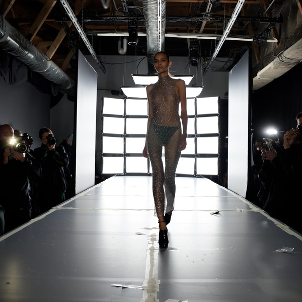 A slender model struts down a glossy silver runway, clad in a sheer, body-hugging ensemble that shimmers with fine metallic threads, every curve outlined like liquid mercury. Her black patent stiletto heels catch soft motion blur mid-step, contrasting sharply with the reflective floor beneath a grid of harsh white LED panels that cast sharp reflections and create delicate, blown-out highlights in the corners. Dark figures clutch cameras in the shadows beside the runway, flashes sporadically popping, while the industrial ceiling reveals exposed trusses and cabling. The runway’s taped sight-lines and slight scuff marks add gritty verité, textured by grainy sensor noise visible in the dimmer spots. Shot from a front-row diagonal, the frame is cropped tightly with a subtle horizon roll and faint hand-shake, capturing the raw pulse of high fashion caught candidly—contemporary runway snapshot, shot on iPhone