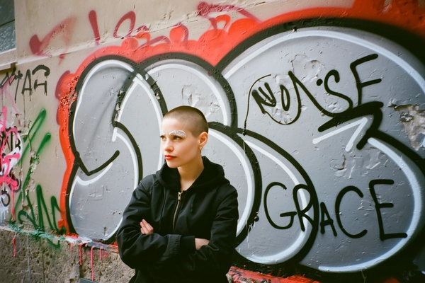 A candid street photo capturing a woman with a buzzcut and sparkling rhinestone eyebrows standing with folded arms. Her calm, defiant expression aligns with the layered graffiti stencils behind her that prominently display the large, stylized text arranged in short lines:
"NOISE"
"GRACE"
The urban wall features overlapping stencil graffiti layers with realistic spray paint drips and weathered textures. Natural daylight softly illuminates the scene, highlighting the textures of peeling paint and rough concrete walls. The framing is spontaneous and slightly off-center with a casual eye-level perspective typical of handheld iPhone photography, conveying an authentic street documentary vibe.