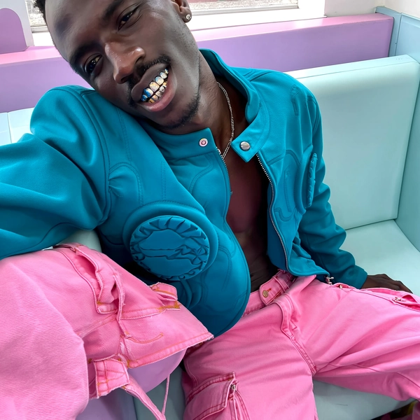 A candid, spontaneous snapshot of a man with a deep skin tone and subtly gleaming golden teeth seated casually in a pastel diner booth with soft, ambient daylight filtering through. He wears a sculptural jacket with distinctive foam texture in bright teal paired with candy pink cargo trousers featuring hardware details, the fabrics showing realistic grain and folds. The pastel environment adds a gentle color contrast, while natural lighting highlights skin texture and the metallic sheen of his teeth. The composition is creatively tilted, capturing a relaxed, unaware pose that feels genuinely spontaneous, characteristic of intimate iPhone photography.