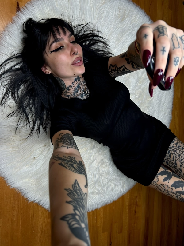 A tattooed young woman lies casually on a circular white fluffy rug atop a warm wooden floor, captured from a bird's-eye view as she holds her phone above for a casual selfie. Her messy black hair fans out naturally around her head, creating a dynamic radial frame. She wears a tight black ribbed short-sleeve dress, showing heavily inked arms, hands, neck, and thighs with bold blackwork floral and abstract tattoos. Her skin texture is soft and detailed under diffused, cinematic lighting that highlights tattoo ink depth and subtle pores. Winged eyeliner and a silver septum piercing accentuate her relaxed expression. Long almond-shaped deep burgundy nails rest lightly on the phone. The composition feels intimate and moody, with casual framing and authentic grunge atmosphere.