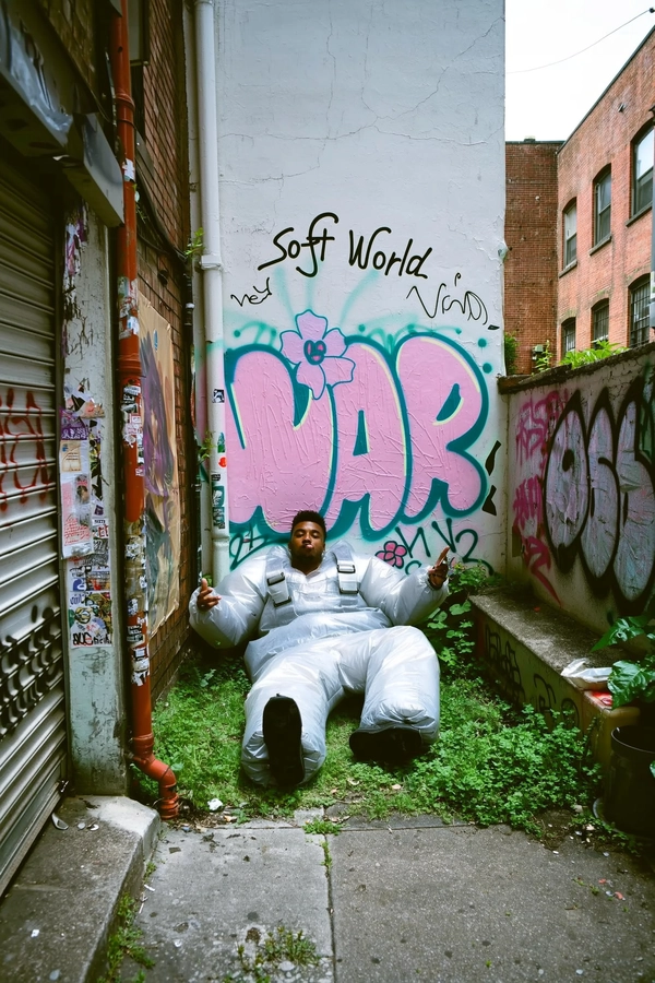 A candid, spontaneous iPhone-style photo capturing a young man lounging nonchalantly in a tiny urban garden next to a graffiti-covered wall. He wears oversized inflatable pants and a translucent plastic harness jacket, his silhouette absurdly contrasted by the soft pastel graffiti behind him, which prominently features large stylized text reading:

"Soft World"
"War"

with delicate flower shapes subtly embedded within the spray paint. The setting is an intimate, slightly overgrown concrete garden nook surrounded by weathered brick walls layered with peeling posters and graffiti tags. Natural daylight filters softly through surrounding buildings, highlighting the matte textures of the inflated fabric and the chalky pastel graffiti. The framing is casually tilted, eye-level, and slightly off-center, emphasizing spontaneous urban authenticity. Visible cracked concrete, subtle rust stains on metal piping, and faint grime details add tactile roughness to the scene. The overall aesthetic evokes a documentary street photo mood with vibrant yet muted tones, true to candid iPhone photographic qualities and street art culture.