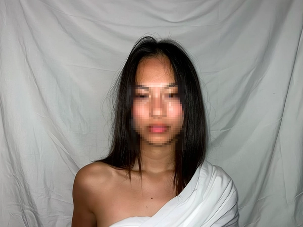 Casual candid portrait of a woman standing against a wrinkled white fabric backdrop, wearing a thin white sheet draped over bare shoulders. Her hair is flat and natural, with realistic, slightly tousled strands. The face is intentionally pixelated to maintain anonymity and add mystery. The scene is lit with raw, direct flash light, producing realistic shadows and highlights typical of spontaneous iPhone photography. Textures of the thin fabric and hair are clearly visible. The framing is straightforward but casually imperfect, suggesting a natural, unposed moment.