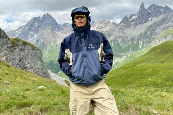 A casual, authentically candid photo spontaneously captured with an iPhone, featuring an individual standing relaxed against a backdrop of jagged mountain ridges and grassy alpine meadows. The person is dressed in a two-tone navy blue and beige Arc'teryx jacket known for its technical design and durability, hooded with the adjustable cords loosely tied, and showcasing weather-resistant fabric vividly creased and softly worn from use. Complementing the jacket are loose-fitting beige cargo pants with natural fabric folds and subtle dirt marks. The individual wears sleek yellow-tinted reflective sports sunglasses, with the hood casually pulled up slightly obscuring the head, and a neutral, composed facial expression. Softly diffused daylight filters through partial clouds, realistically illuminating fabric textures, rugged rock details, and lush grass blades surrounding the figure, capturing authentic mountainous outdoor ambience. The slightly tilted framing emulates spontaneous iPhone photography style with a subtle editorial vibe, emphasizing genuine, trendy technical outdoor aesthetic and casual intimacy typical of gorpcore fashion.