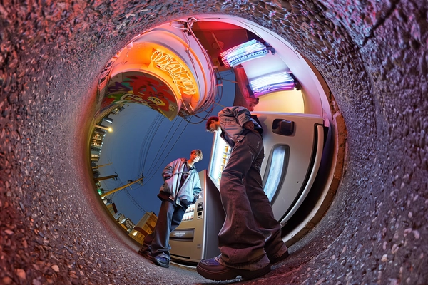 A fashionably spontaneous 360-degree candid shot captured from ground level with a spherical camera paired with an iPhone, focused upward creating a tunnel-like circular distortion. Two models stand casually leaning against a sleek urban vending machine positioned in a narrow alley. They wear futuristic cyber-chic windbreakers featuring lightweight technical fabrics in muted metallic and deep matte tones layered with translucent, layered visors that subtly reflect neon glows. Their relaxed postures and natural interactions reveal effortless modern style, with tapered streetwear pants and minimalist sneakers showing authentic textured wear, complemented by subtly tousled hair and nuanced skin textures with natural imperfections. Overhead, dense cables, vibrant layered graffiti walls, and glowing neon signs spiral upward into the night sky, bathed in cool urban neon blues and magentas. Soft, atmospheric ambient neon illumination casts dynamic yet realistic shadows on gritty concrete and wet pavement textures below, emphasizing tactile details. The immersive upward-facing spherical framing generates a striking circular composition with stylishly distorted architectural lines and environmental elements, authentically replicating spontaneous candid 360-degree iPhone photography from a low central ground perspective, evoking a raw, cyber-street aesthetic with genuine casual elegance.