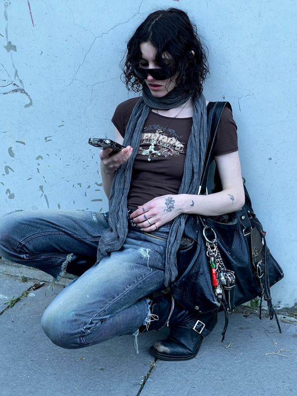 A painfully thin young woman slouches against a cracked, graffiti-speckled concrete wall in a mid-close iPhone shot with a slight tilt and uneven framing that cuts just below her knees. The ambient light is flat and bluish, soft natural daylight casting subtle shadows that highlight faint bruised undertones beneath her eyes. She wears a threadbare chocolate brown graphic tee clinging tightly to her collarbones, unevenly tucked into low-rise distressed skinny jeans mottled with faded patches and fraying just above one knee. Over it, a shrunken black denim jacket with worn seams and missing buttons hangs loosely. A long, gray scarf is casually wrapped and dropped from her neck, moving lightly with her breath. Black leather boots with scuffed toes and worn buckles anchor her look, and an oversized weathered black leather satchel, adorned with metal keychains and charms, droops heavily at her side. 

Her dark curly hair falls messily over her face, partially hiding a pair of large black sunglasses perched precariously on the bridge of her nose. She looks down at her hand, fingers casually holding a cracked smartphone, eyes unfocused, as if lost in a text or thought. Tattoos peek subtly on both hands with rough linework. Her pale skin reveals natural pores, faint redness, and some subtle dryness, capturing raw intimacy. The entire scene feels unfiltered, immediate and spontaneous, like a candid snapshot of haunted indie sleaze youth caught in a quiet urban moment—dark, fragile, and undeniably real.