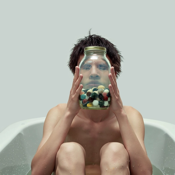 A surreal minimalist portrait showcasing a shirtless model seated in an empty bathtub against a smooth pale gray background devoid of distractions. They hold a transparent mason jar completely filled with murky water and assorted marbles, positioned directly in front of their face, fully covering and drastically warping their features into primitive, abstract shapes evocative of early 2000s editorial absurdity. Muted, softly pastel color grading and authentic soft daylight softly illuminate the scene, capturing realistic skin texture and subtle water refractions through the curved glass surface. The composition uses spontaneous informal framing and a natural candid angle, emphasizing raw intimacy and surreal distortion. Styling is pared down to bare skin, focusing on natural hair strands and subtle makeup, embodying textural personalization through the contrast of smooth skin and irregular glass distortions. The overall aesthetic channels contemplative authenticity and surreal minimalism reminiscent of a spontaneous iPhone photograph.