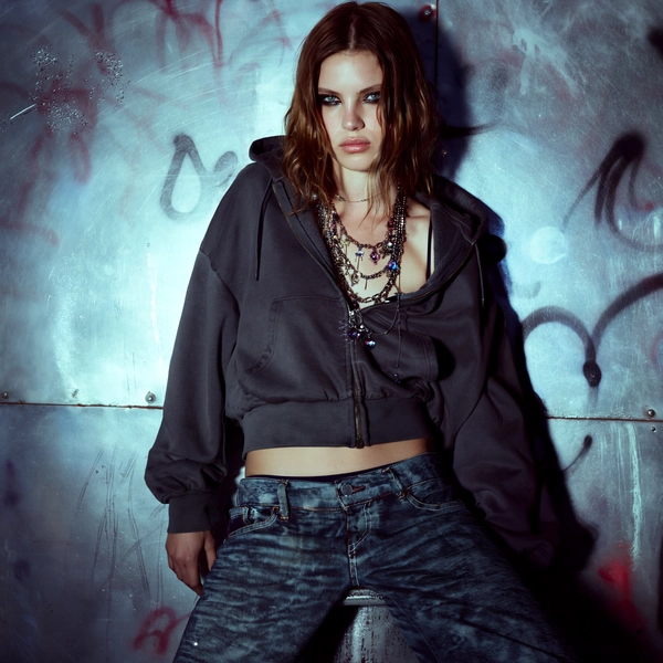 A young woman stands poised against a graffitied metal wall, the scene saturated with the cold shimmer of fractured neon blues and subdued iron greys. Her charcoal grey hoodie, unzipped to frame meticulously styled layered necklaces, reveals low-rise jeans with a raw, lived-in denim texture that invites tactile attention. Smudged black eyeliner has a deliberate, smoky edge that contrasts with her glistening, red-rimmed eyes—there’s a tension held in her steady, unyielding gaze beneath tousled, natural hair accented by a faint dewy glow.

Her stance leans lightly into the graffiti-harboring steel, sculptural in its relaxed gravitas, shoulders tilted against the rugged backdrop, arms loosely crossed yet fluid, capturing a moment suspended between defiance and vulnerability. The interplay of digital flash scuffs the scene with a hazy veil, softening the hard chrome textures while accentuating the sheen of her hoodie’s brushed cotton weave, the rough metal’s peeling paint, and the layered jewelry that catches glinting rim lights.

A stark studio setting, lost in shadow but punctuated by bursts of color from neon gels, melds with the gritty urban canvas—a cinematic tableau where cold light fractures over skin pores and the subtle fingerprints smeared across her metal surroundings. Framed from a low angle with a 50mm lens, the shot compresses the wall’s sprawling graffiti into an abstract wash behind her, edges softly blurred to draw focus on her direct eyes and layered texture-rich elements.

Digitally processed with a muted color grade ramping from steely cyan to subtle charcoal, this image channels the spirit of gritty, neo-urban fashion editorials, emphasizing hyper-real fabric weaves and skin detail with a hint of surreal flash backdrop—high-fashion editorial, hyper-real texture fidelity