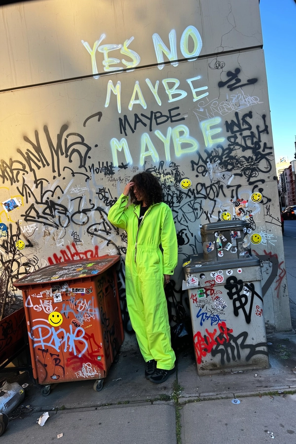 A candid urban street photograph of a woman wearing a vibrant lime green utility suit standing casually in front of a heavily graffiti-tagged concrete wall. The wall features the repeated phrases arranged visually as a looping echo reading:
"YES NO"
"MAYBE"
"YES NO"
with some of the graffiti glowing faintly as if illuminated by subtle neon paint. Surrounding her are cluttered urban elements—rusty trash bins and weathered electrical boxes—also covered with climbing graffiti tags and layered slap stickers including iconic smiley faces and cartoon characters. The natural late afternoon daylight casts soft shadows enhancing the rough textures of peeling paint, cracked concrete, and peeling sticker edges. The framing is slightly tilted with an eye-level, spontaneous angle typical of an iPhone photo capturing raw street authenticity. The overall image conveys gritty, vivid urban realism, consistent with documentary street photography aesthetics.