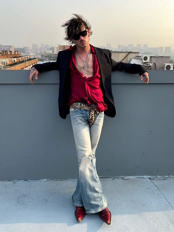 A young man leans casually against a metal railing on a high-rise rooftop, shot mid-frame with an off-center crop cutting just below his knees. The hazy late afternoon sky casts flat, natural light over concrete underfoot, shadows soft and diffused. Behind him, distant city buildings and HVAC units blur softly, their outlines punctuating the urban skyline. A breeze lifts and billows the edge of his deep red silk shirt, slightly unbuttoned to reveal chunky silver chains and faint chest skin texture. He wears oversized retro black sunglasses that obscure his expression, and a fitted black blazer with worn creases hugging his shoulders.

His light-washed flared jeans sit low on his hips, cinched with a vintage leopard-print scarf belt, the fabric faded, wrinkled, and worn. Dark red pointy cowboy boots peek beneath, scuffed and creased. His rocker-style mullet hair is messy, with oily roots and uneven lengths tousled by the breeze. Exposed hands rest loosely on the railing, skin showing natural pores, redness, and faded knuckle tattoos. His head tilts away from camera, posture relaxed but dramatic, captured with unpolished immediacy typical of an iPhone photo—grainy, candid, and thick with gritty glam rock revival energy.