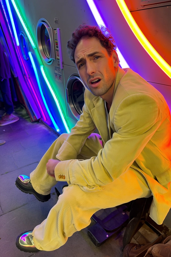 A spontaneous candid snapshot of a man with narrow eyes and high cheekbones sitting casually in a laundromat illuminated by an array of colorful neon tubes, casting soft multicolored glows on his lemon-yellow corduroy suit. The suit's textured fabric and exaggerated cuffs are vividly detailed alongside his platform-soled patent shoes catching subtle reflections. Natural skin textures and a relaxed, unaware expression add genuine authenticity. The lighting balances ambient neon hues with natural shadows, typical of an iPhone photo taken indoors at twilight. The composition is informally framed with a slight tilt, evoking a casual, spontaneous moment of urban life.
