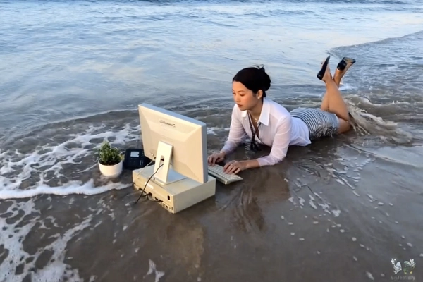 With waves lapping rhythmically against the shore, a woman lies prone in shallow ocean water, her white blouse and striped pencil skirt crisp despite the gentle surf swirling around her. She calmly types on an old beige keyboard, the vintage CRT monitor and stacked PC tower perched precariously in front, half-submerged and glistening under the warm evening light. Nearby, a potted plant and a wired office phone complete the surreal desktop arrangement on the wet sand. The photo has a grainy quality, suggesting it may be a still from a video or taken with an older digital camera. Her dark hair is pinned neatly in a tidy bun, while her serene expression remains oddly composed amidst the shimmering reflections. Quick snapshots create a soft-focus effect at the edges, and muted colors swirl into a nostalgic haze, capturing an absurd twist on remote work culture. The faint breeze carries a salty tang as waves ripple gently around her wrists, merging the rigid formality of office life with beachside tranquility—early-2000s digicam snapshot, grainy quality.