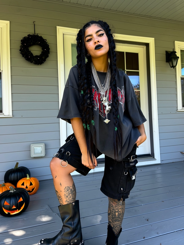 A bold young woman stands casually on a grey wooden suburban porch decked with Halloween decor—black wreaths, carved pumpkins, and a windowed front door visible behind. She's captured in a mid-close shot from knees up with relaxed posture, looking just past the camera. Her long dark hair is braided in thick plaits with subtle green accents, and her skin shows natural texture with visible pores and faint blemishes on her arms. She wears an oversized black nu-metal band tee with distressed red and white lettering, paired with black cargo knee-length shorts adorned with D-rings and metal loops. Her legs are tattooed with detailed black ink visible under soft natural daylight. Heavy layered silver chains hang around her neck. Platform black combat boots with silver studs peek from frame bottom. Bold sharp eyeliner and matte black lipstick accent her expressive face. The lighting is natural with soft shadows and gentle highlights, producing an intimate, candid feel.