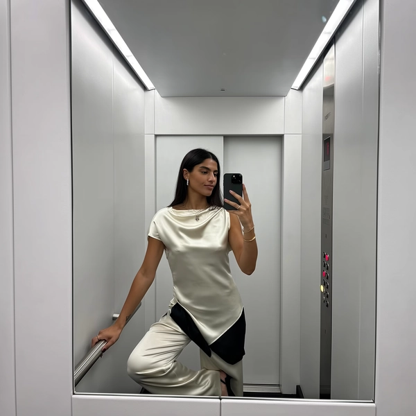 In the clinical hush of a stark white elevator, her slinky asymmetric silk top glides softly over fluid trousers, the satin sheen catching the cool LED strips above with a quiet gleam. One leg bends with casual poise, the black button panel punctuating the purity of the space behind her. Her neutral expression lifts slightly toward the screen as the phone rests just below her face, fingertips poised mid-press; the slight barrel effect wraps the minimal surroundings, while a faint smudge laces the corner of the frame, lending texture to her smooth skin and the subtle weave of her outfit. The light plays cool and graphic against the monochrome calm, encapsulating that effortless urban elegance—a fleeting moment of poised ease, waiting between floors. —elevator mirror capture, urban chic on iPhone