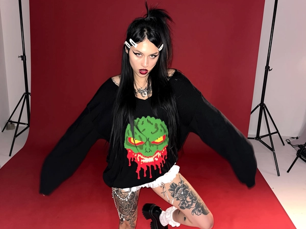A fierce young woman stands against a deep blood-red studio backdrop, shown from mid-thigh up in a casual, slightly angled mid-close shot. Her long jet-black hair is styled in a messy half-up ponytail, adorned with small white clips, with natural flyaways visible. She wears an oversized black sweater featuring a vivid graphic of a melting monstrous green face with yellow eyes and dripping red accents resembling flames, the fabric textured with gentle wrinkles. Below, white frilly bloomers peek out subtly. Her tattooed legs display large, detailed black floral and demonic motifs, layered with loosely hanging metal chains as accessories. Chunky black platform shoes with white frilly socks complete her look. She wears bold winged eyeliner and deep lipstick with natural skin texture visible on her pale face. Lighting is high-contrast studio but softened to mimic iPhone warmth, lending an eerie yet playful atmosphere. The composition feels relaxed and spontaneous, with slight tilt and imperfect framing.