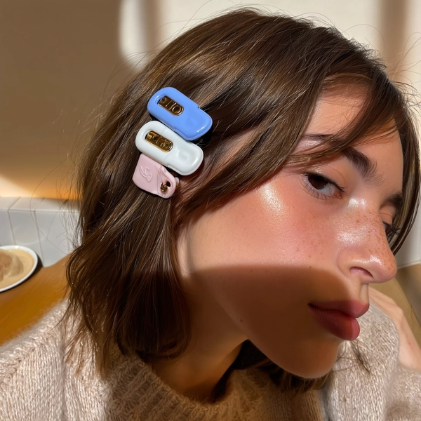 Pastel blue and white barrettes cluster playfully on one side of shoulder-length brown hair, their smooth sheen catching the gentle café window glow. The girl's flawless skin shimmers with a glassy glow, dewy and fresh, as loose strands skim her freckled temple. A slight high-angle stretch captures her calm stare, framed by softened shadows and the faint fuzz of a knit collar brushing her jawline. The background blurs softly, hinting at a cozy cafe nook; a stray coffee cup rim and tiled backsplash peek out, grounding this candid moment while subtle reflections shimmer on the clips' surfaces. The casual tilt of her head and the warm light make this intimate scene feel effortlessly authentic—authentic iPhone close-up, statement hair-clip focus