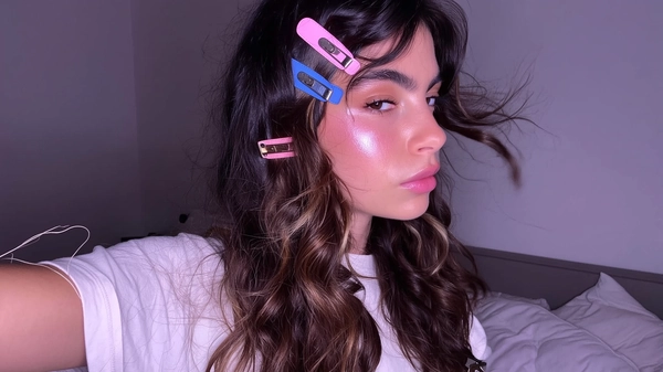 Mixed pink and blue Y2K-style clips scatter whimsically through glossy wavy brunette hair, the pastel sheen catching soft, moody bedroom light that skims her calm face with a delicate pout. Loose tendrils frame her cheeks, each clip glinting with a slight worn texture, while her barely-there makeup and glass-skin glow add a fresh, effortless vibe. The shot dips gently into a casual arm-extended angle, bed pillows forming a blurred but cozy backdrop. A stray thread on her cotton tee peeks near the frame, while skin texture and fly-away strands sing quiet authenticity—authentic iPhone close-up, statement hair-clip focus