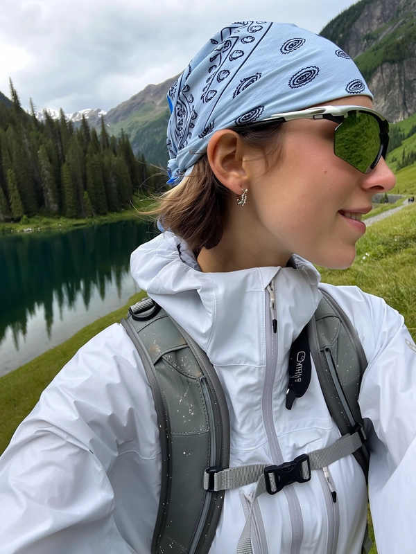 A spontaneous, candid close-up portrait captured with an iPhone, featuring a young woman hiking in the mountains. She wears a crisp white hooded windbreaker jacket with a high collar, made of smooth weather-resistant fabric showing subtle creases and realistic wear. Gray hiking backpack straps cross her chest securely, textured with slight fabric folds and natural dirt marks from outdoor use. Her head is wrapped in a light blue bandana patterned with black paisley designs, tied neatly to reveal soft natural hair underneath. Futuristic wraparound mirrored sunglasses reflect the serene alpine lake and coniferous forest behind her. The woman's skin appears fresh and natural with minimal makeup, small metallic earrings visible, enhancing the functional streetwear aesthetic. The background reveals a calm mountain lake surrounded by dense evergreen trees and distant peaks under a gentle, overcast sky. Soft, diffused daylight illuminates the scene, bringing out authentic textures in the jacket fabric, backpack, and skin details. The framing is a tight, slightly tilted close-up angle typical of candid, stylish iPhone outdoor photography, imparting a peaceful, utilitarian mood infused with natural beauty and sleek technical fashion.