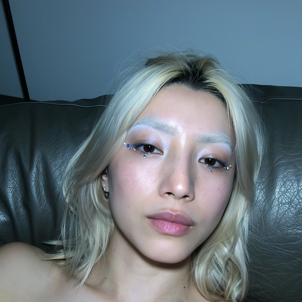 A natural close-up iPhone selfie of a young woman with platinum hair styled casually, featuring naturally bleached eyebrows that softly blend into her fair skin. Her eyes are adorned with subtle icy shimmer eyeshadow accented by delicate crystal embellishments, capturing a refined yet effortless look. She lies on a leather couch, the dark textured material visible behind her, while the dim room casts cool, soft shadows that gently highlight her facial features and makeup's subtle glossiness. Her expression is calm and neutral, engaging the camera directly with a spontaneous vibe. The overall scene maintains a minimalist and neutral color palette, with the leather couch adding understated texture, creating a candid, stylish, and contemporary portrait typical of authentic iPhone selfies.