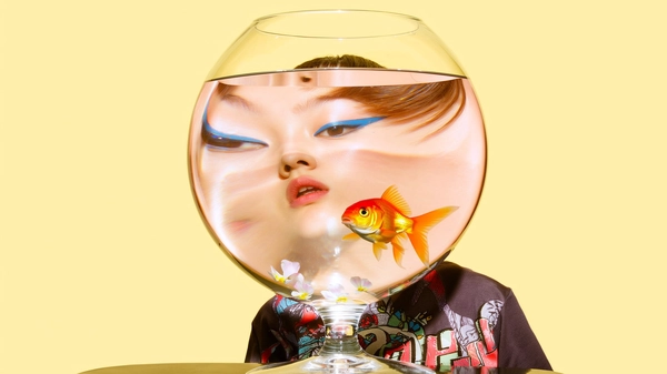 A surreal, minimalist portrait of a young person with short bangs and striking blue eyeliner, styled in a casual playful outfit featuring a relaxed graphic tee and creatively mixed accessories. They place a square transparent fishbowl, entirely filled with clear water, directly over their head so it fully covers and obscures the face. Inside the fishbowl, a fake goldfish and scattered delicate flower petals float, their presence enhancing surreal distortions. The water refraction shifts and elongates the facial features upward unnaturally. The scene is set against a smooth softly pastel yellow background, illuminated by bright yellow natural daylight that casts gentle reflections and shadows, emphasizing the playful absurdity. Realistic skin texture, subtle makeup nuances, and detailed hair strands are visible through the glass. The spontaneous, intimate framing uses casual angles to center the fishbowl, producing a strikingly surreal yet authentic image evocative of candid iPhone photography.