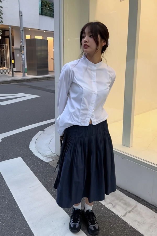 A casually candid iPhone photo of a young woman patiently waiting at a crosswalk in an urban minimalist setting with Japandi influences. She wears a crisp white high-collar shirt neatly tucked into a long, textured navy pleated skirt, paired with chunky heavyweight sneakers and thin white socks subtly peeking above, embodying the relaxed yet refined Japandi aesthetic. Her posture is upright but relaxed, her natural hair loosely styled with minimal makeup, highlighting subtle skin textures and natural expression. The ambient lighting mimics early evening streetlight glow softly reflecting on a nearby sleek glass panel, casting delicate highlights and diffused shadows on her and the surrounding concrete pavement. The background includes minimalist urban elements with muted tones—simple gray concrete, faint street markings, and a few minimalistic metallic structures suggesting contemporary city architecture. The framing is slightly tilted and intuitively spontaneous, capturing an authentic iPhone snapshot feel with gentle focus shifts enhancing the candid atmosphere and tactile fabric details, natural skin nuances, and subtle reflections, creating a genuine moment of urban Japandi style composition.