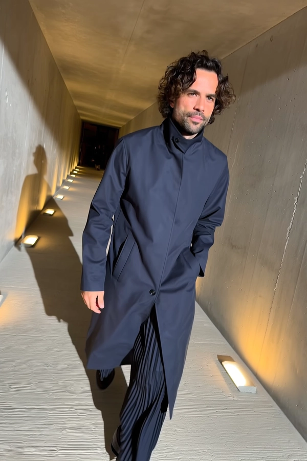 A candid moment of a man walking through a sleek modern concrete hallway, dressed in a matte navy trench coat with a high collar and wide-legged pleated pants. Soft curls frame his face, highlighting defined cheekbones under the ambient glow of floor lights that cast elongated shadows on the textured concrete walls and floor. Natural lighting from the hallway enhances the tactile fabric details and subtle skin textures. The framing is slightly off-center and tilted, emulating a spontaneous iPhone snapshot that feels intimate and effortlessly stylish.